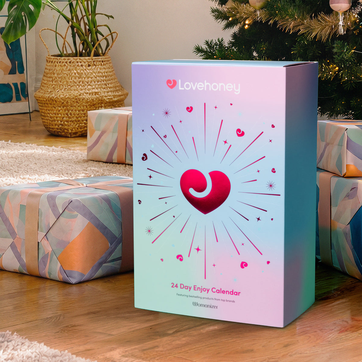 A Lovehoney 24 Day Enjoy Calendar box by Womanizer, styled on a wooden floor next to wrapped Christmas presents and a potted plant, with a Christmas tree in the background.