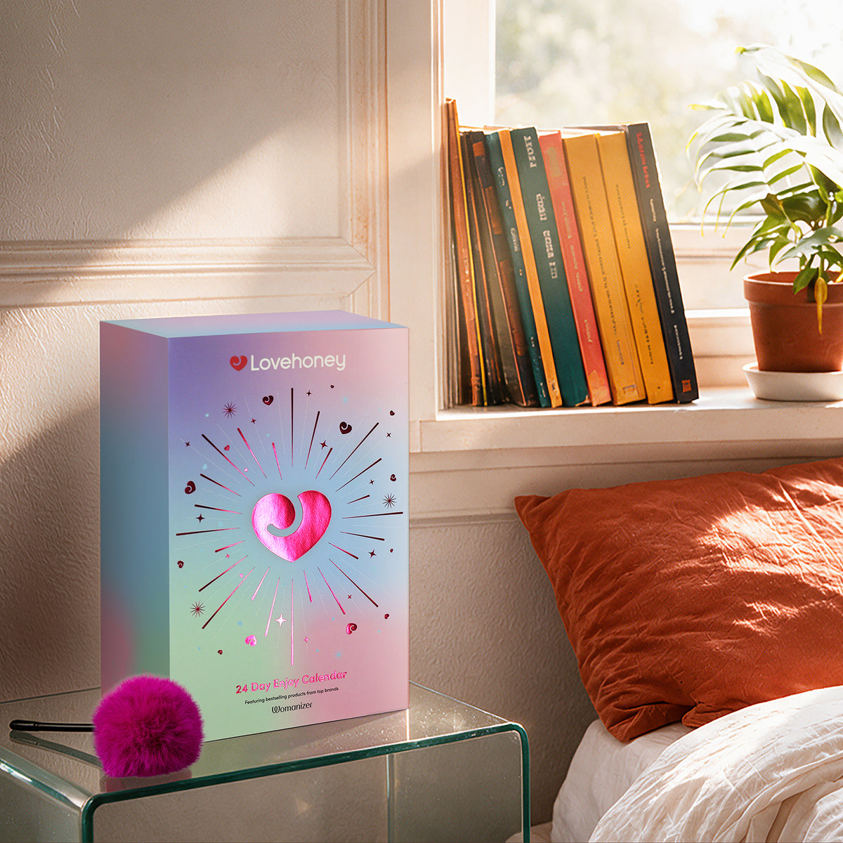 A pastel gradient Lovehoney Valentine's Day gift box sits on a glass side table in a sunlit bedroom, next to a bright pink fluffy pom-pom. The background shows a window with books and a potted plant.