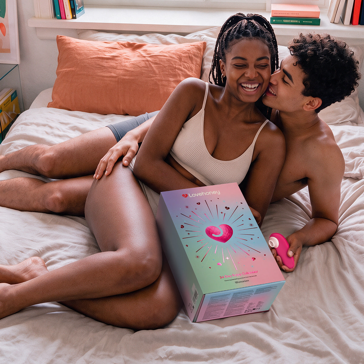 A couple lying on a bed in a bedroom, with the woman holding a pink personal massager and the man kissing her cheek. A large pastel product box labeled 'Lovehoney' sits on the bed in front of them.