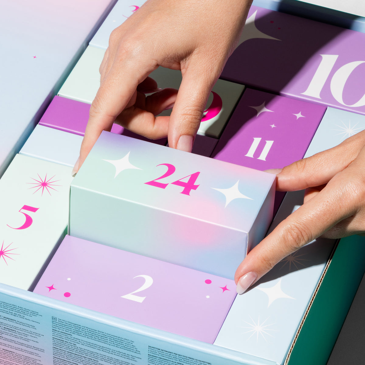 A close-up of hands opening a pastel advent calendar, lifting the numbered 24 box. The calendar has multiple compartments in soft pastel shades of purple, pink, mint green, and white, arranged in a grid.