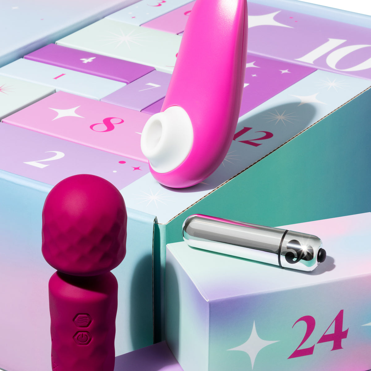 Two intimate wellness devices displayed with a pastel advent calendar box: a bright pink curved wand-style device and a darker magenta textured handheld device, alongside a silver capsule-like item on a branded box marked 24.
