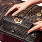 A close-up of a black and gold advent calendar being opened by two hands, with the front door numbered 24 pulled down. Other numbered compartments are visible around it.