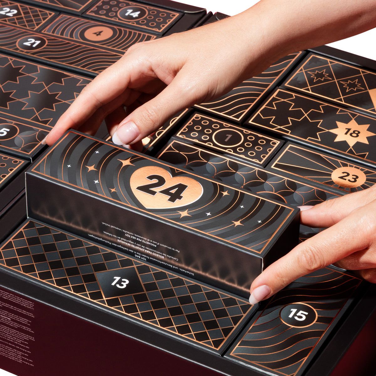 A close-up of a black and gold advent calendar being opened by two hands, with the front door numbered 24 pulled down. Other numbered compartments are visible around it.