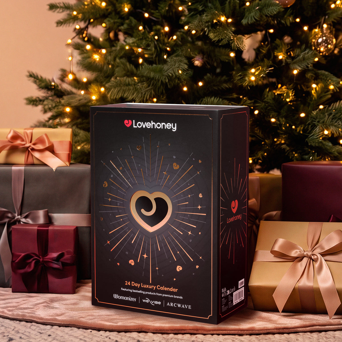 Lovehoney 24 Day Luxury Calendar box displayed on a rug in front of a decorated Christmas tree with wrapped gift boxes around it.