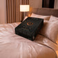 A black rectangular product box with gold and red details sits on a bed with pinkish-beige bedding and pillows in a softly lit bedroom. The box has the Lovehoney brand name visible near the top.