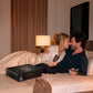 A man and woman lying together on a bed in a warmly lit bedroom, smiling at each other while holding black handheld devices. A black product box with a gold circular logo sits on the bed beside them.