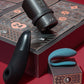 A boxed adult novelty set displayed on a red surface, featuring a black cylindrical device resting on the open box lid, a matte black curved piece in front, and a teal curved piece on a small box marked 1. The packaging has a dark pattern with numbered compartments and copper-toned lines.