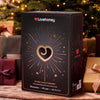 A Lovehoney 24-Day Luxury Advent Calendar box, presented in a festive Christmas setting with wrapped gifts and a decorated tree in the background.