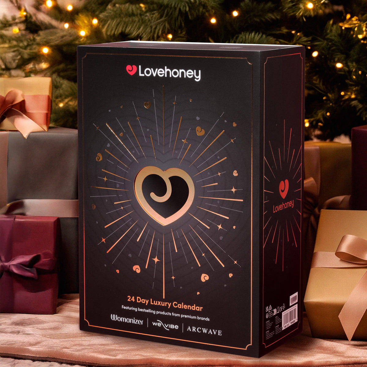 A Lovehoney 24-Day Luxury Advent Calendar box, presented in a festive Christmas setting with wrapped gifts and a decorated tree in the background.