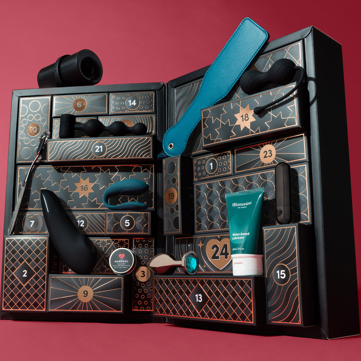An open black advent calendar gift box with numbered compartments, filled with intimate wellness products including a teal strap, black massager, teal tube, and other accessories, set against a solid red background.