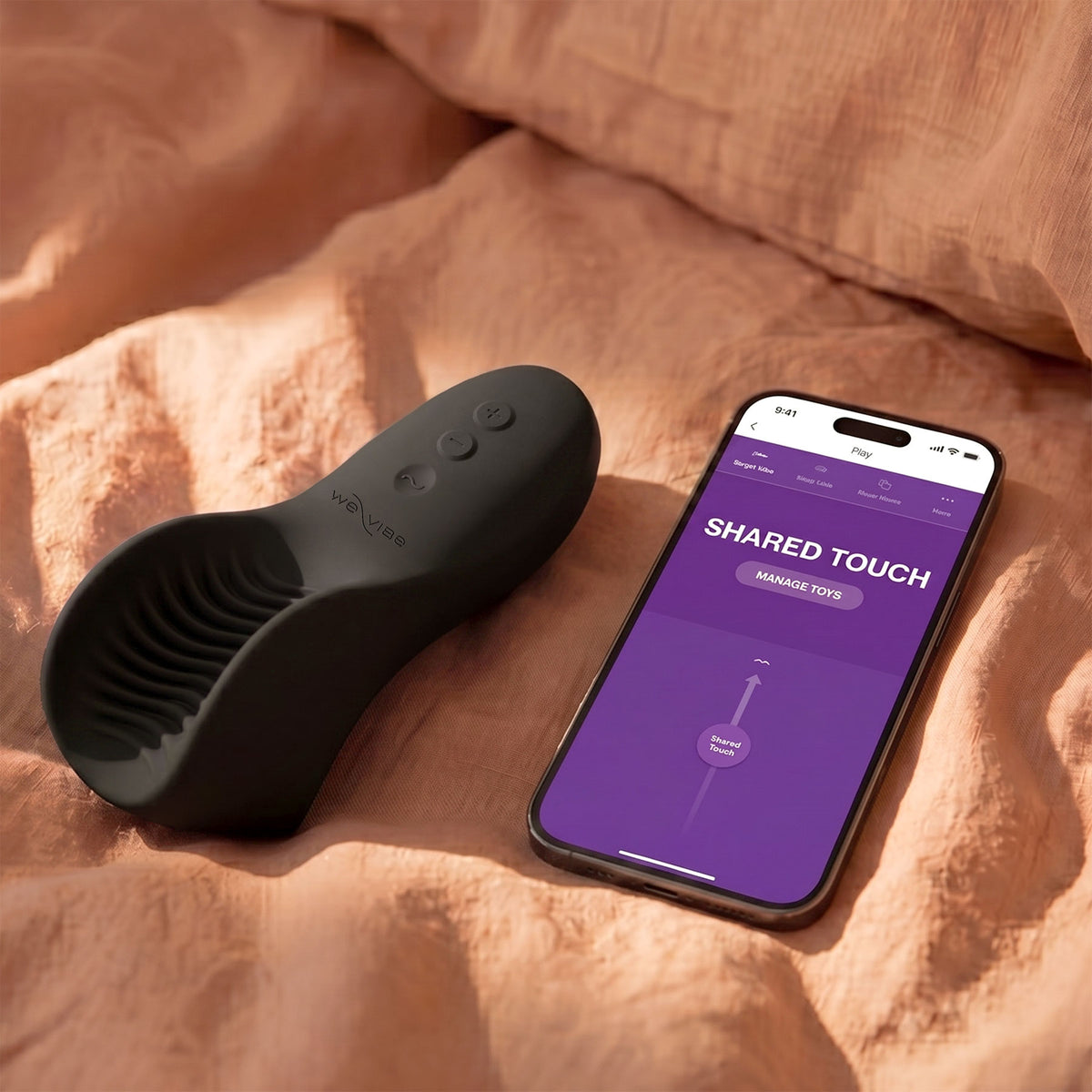 A black personal massager with a ribbed suction-style head rests on peach-colored bedding next to a smartphone displaying a purple app screen that reads 'SHARED TOUCH'.