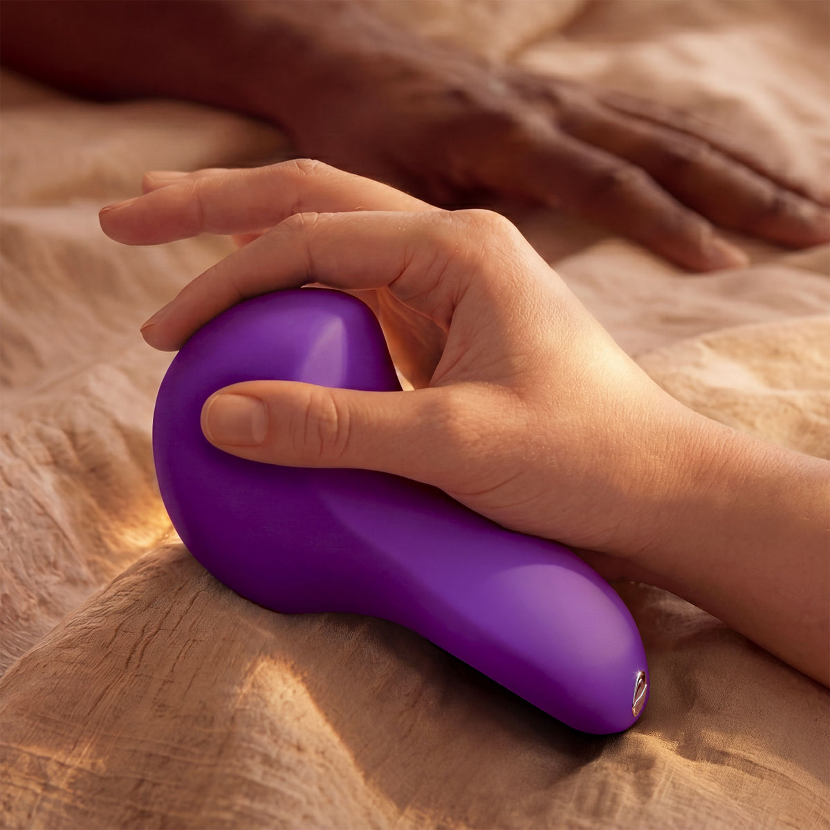 A close-up of a hand holding a purple, teardrop-shaped personal massager on a beige textured blanket, with another hand blurred in the background.